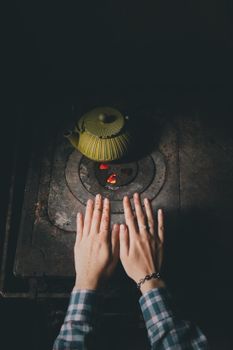 Hands above a vintage stove with a teapot, offering warmth and comfort.