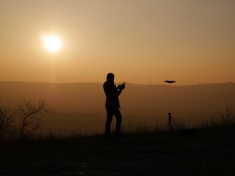 Full body silhouette of anonymous person standing on slope at sunset and using remote control for operating drone