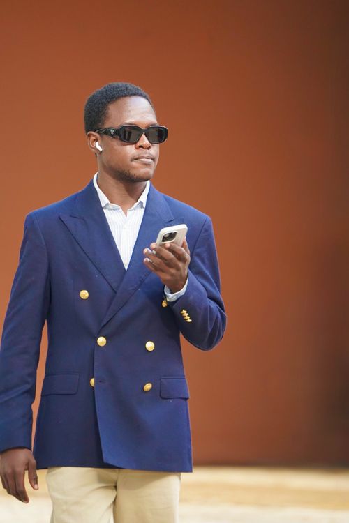 Free Fashionable man wearing sunglasses and a blue blazer walks confidently while holding a smartphone. Stock Photo