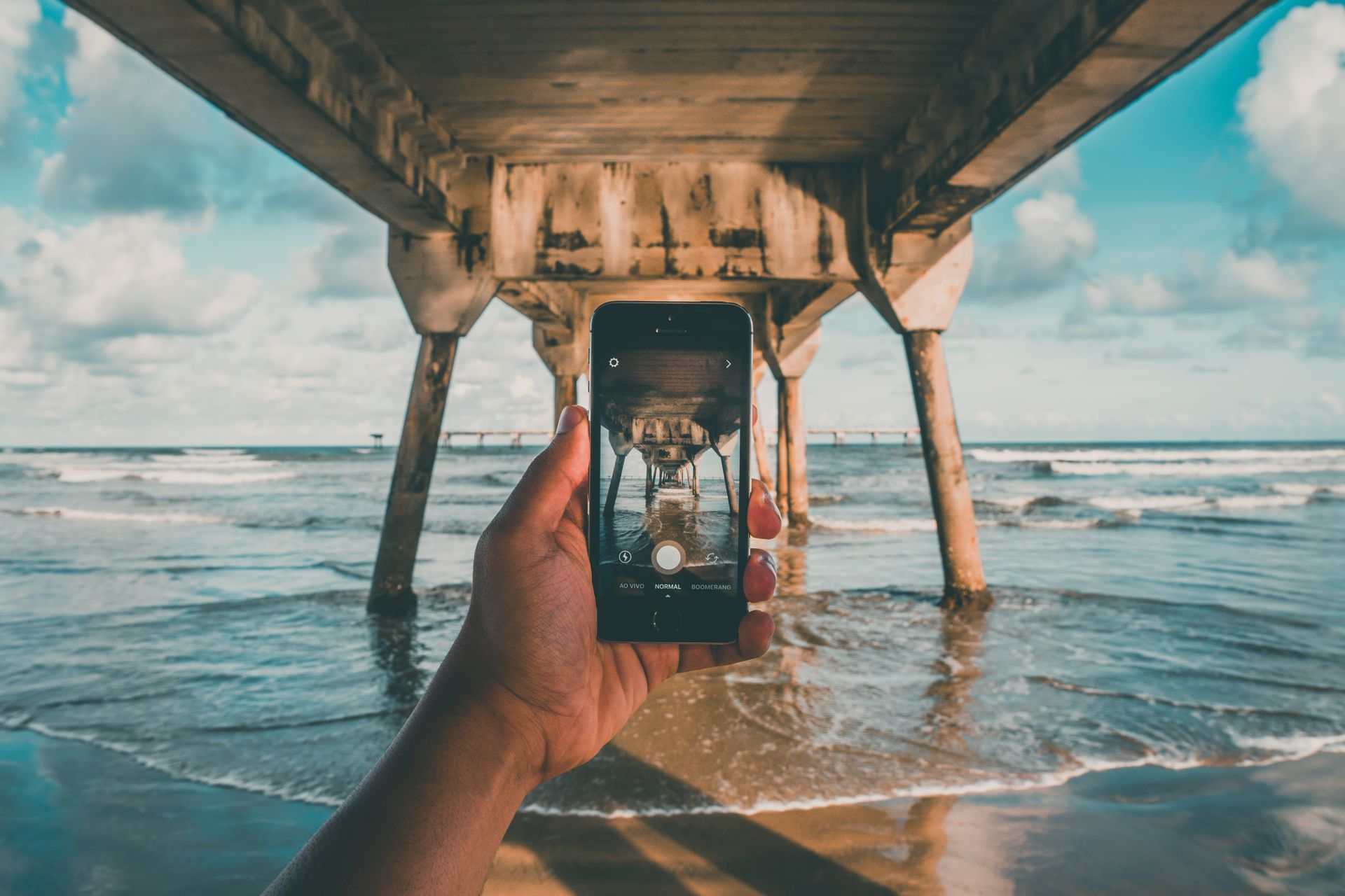 A hand captures a stunning ocean view beneath a pier using a smartphone, emphasizing creativity in mobile photography.