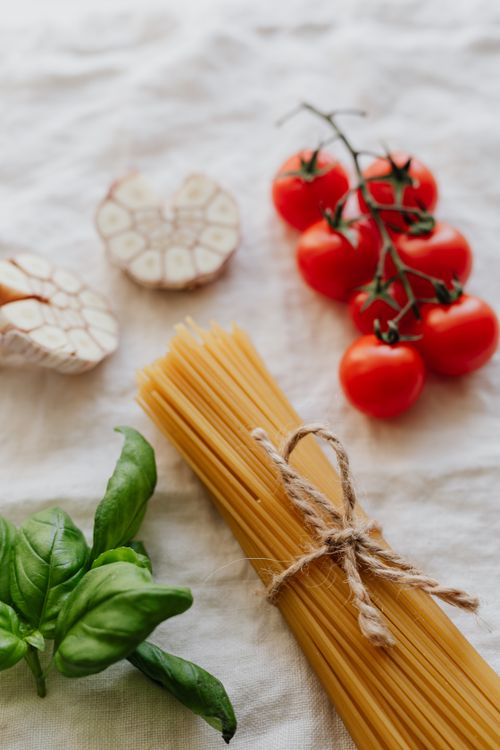 Free High-angle view of fresh ingredients: spaghetti, cherry tomatoes, garlic, and basil on a rustic cloth. Stock Photo