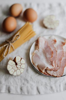 Flat lay of pasta dish ingredients: bacon, eggs, garlic, spaghetti on rustic cloth.