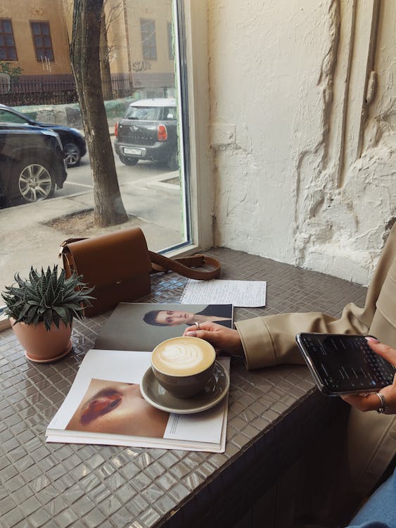 Free Crop of unrecognizable female tourist in casual wear surfing internet and flipping magazine while drinking coffee in sidewalk cafe Stock Photo