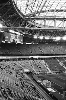 Black and white photo of a modern domed stadium with empty seats and a large screen.