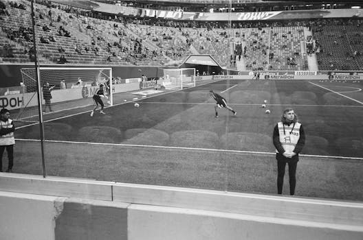 Black and white photo capturing a live football match with players in action and spectators in the stadium.