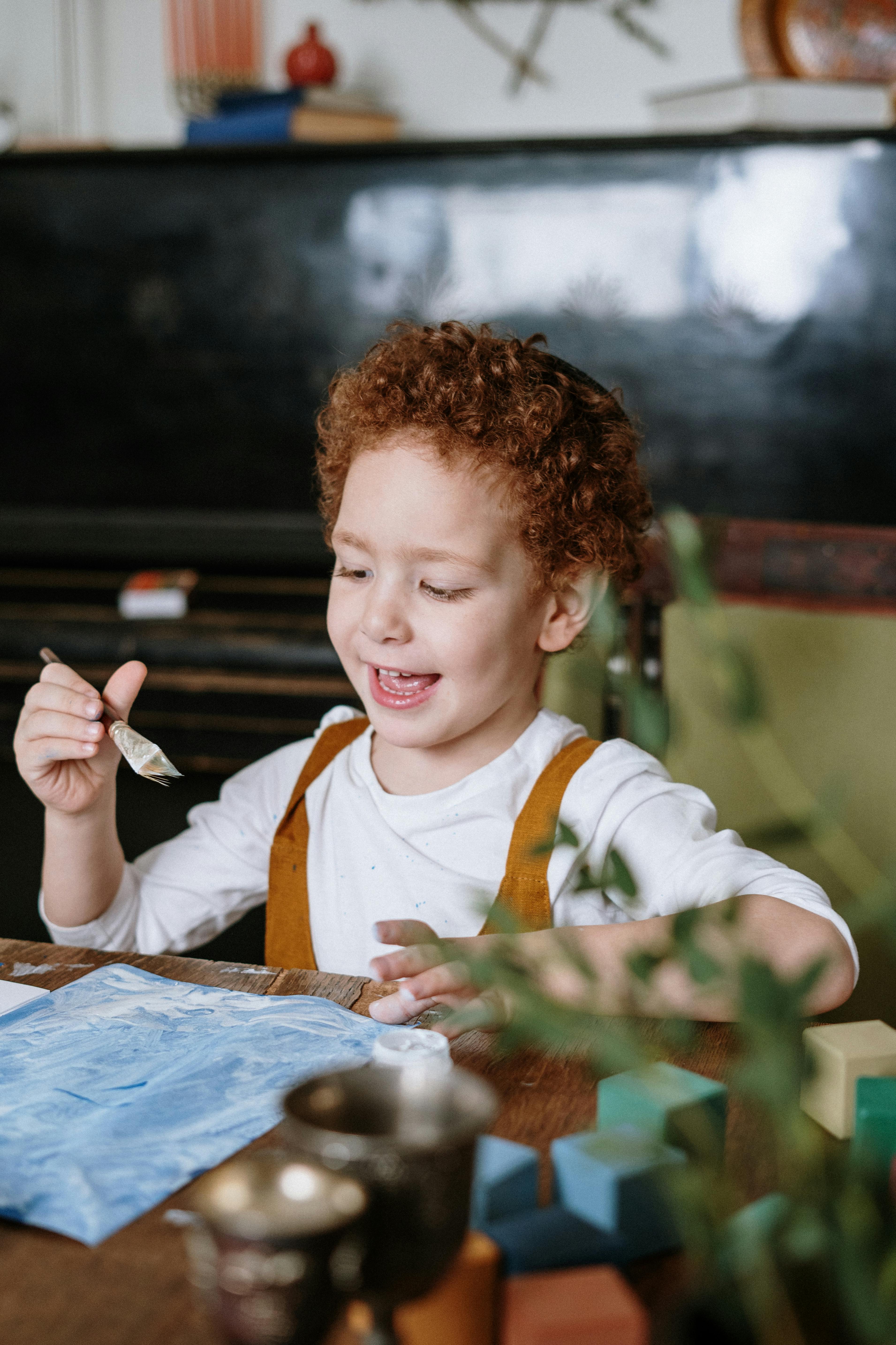 一个开朗的孩子，顶着一头卷曲的红发，正在室内开心地画画，尽情表达着喜悦与创造力