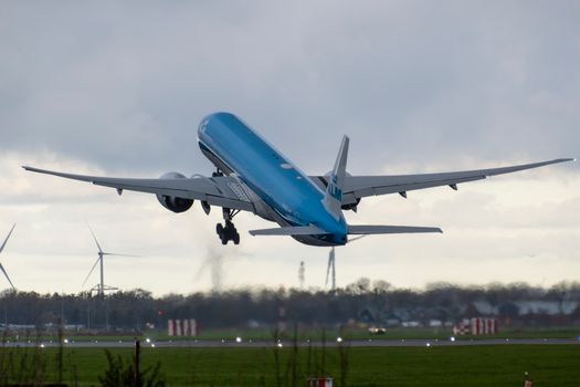 A blue airplane taking off with wind turbines in the distance on a cloudy day.