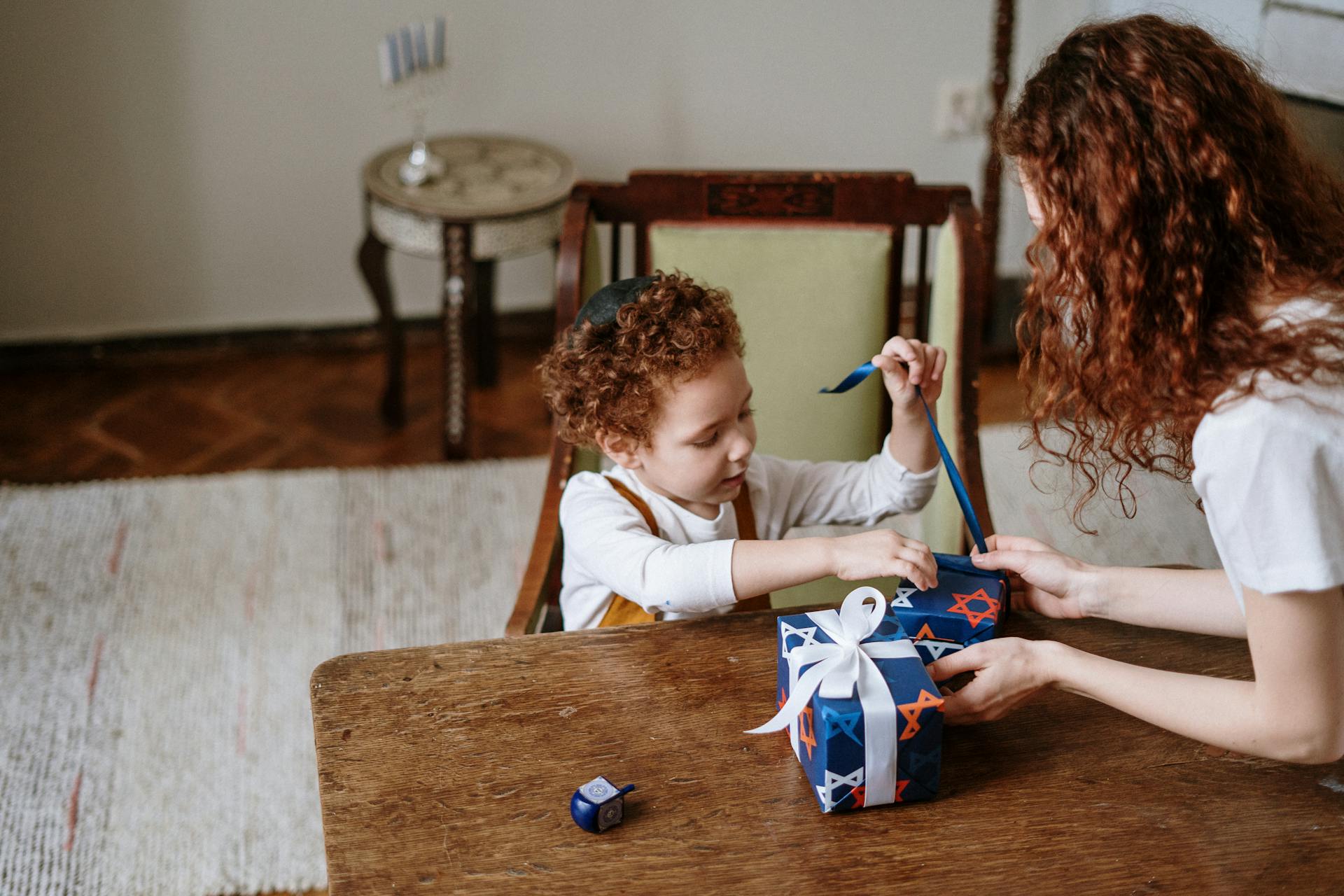 A mother and child joyfully wrapping a Hanukkah gift at home with a dreidel on the table.