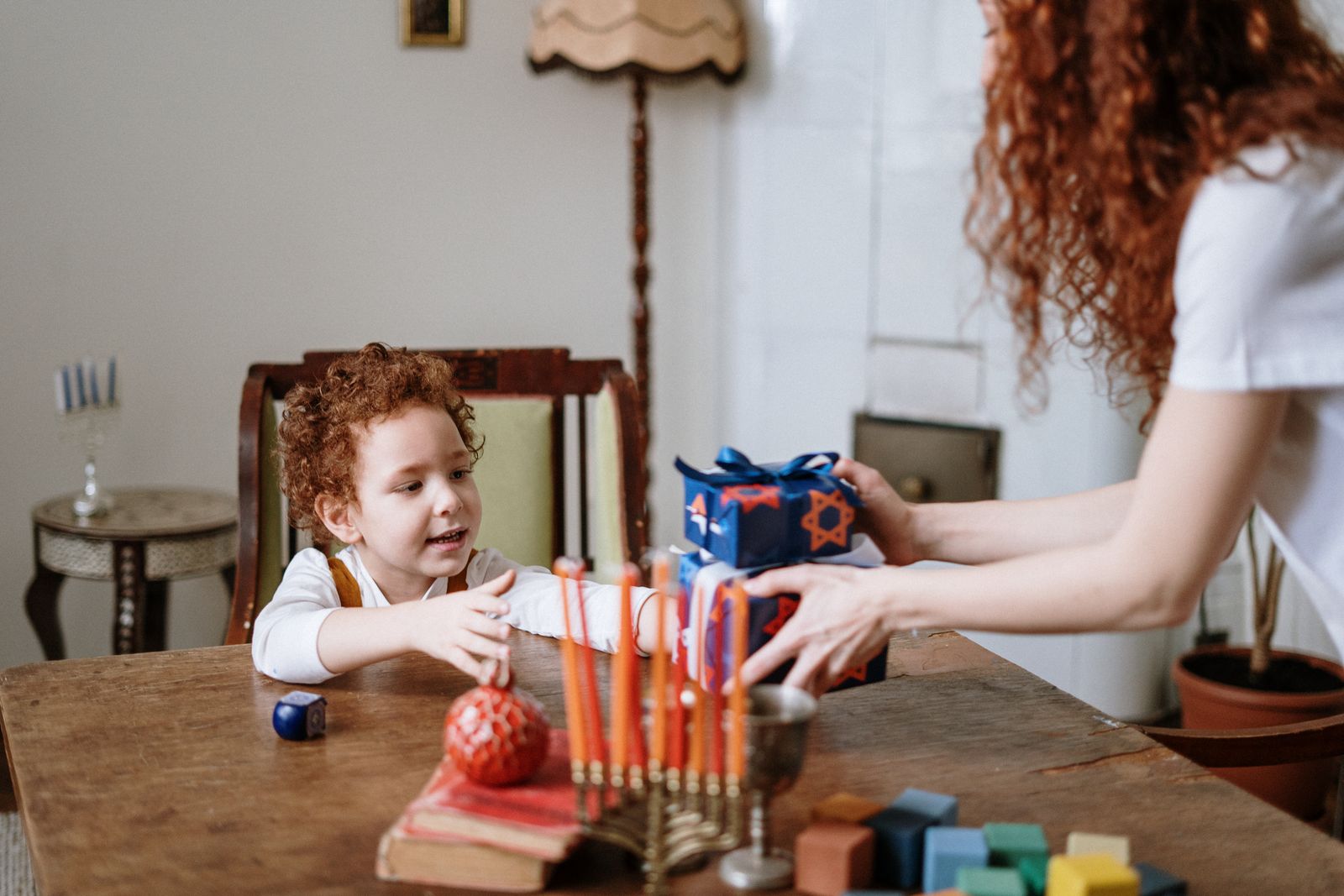 Mother handing a wrapped Chanukah gift to her young son across a table with a menorah