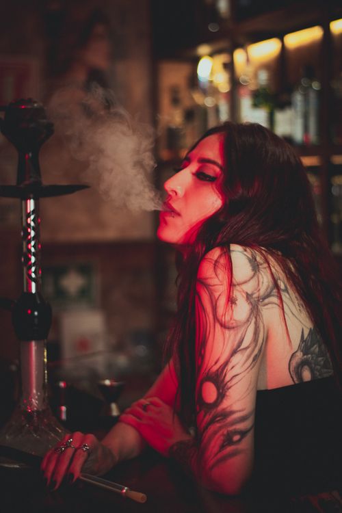 Free A young woman with tattoos relaxes in a bar, exhaling hookah smoke under warm lighting. Stock Photo