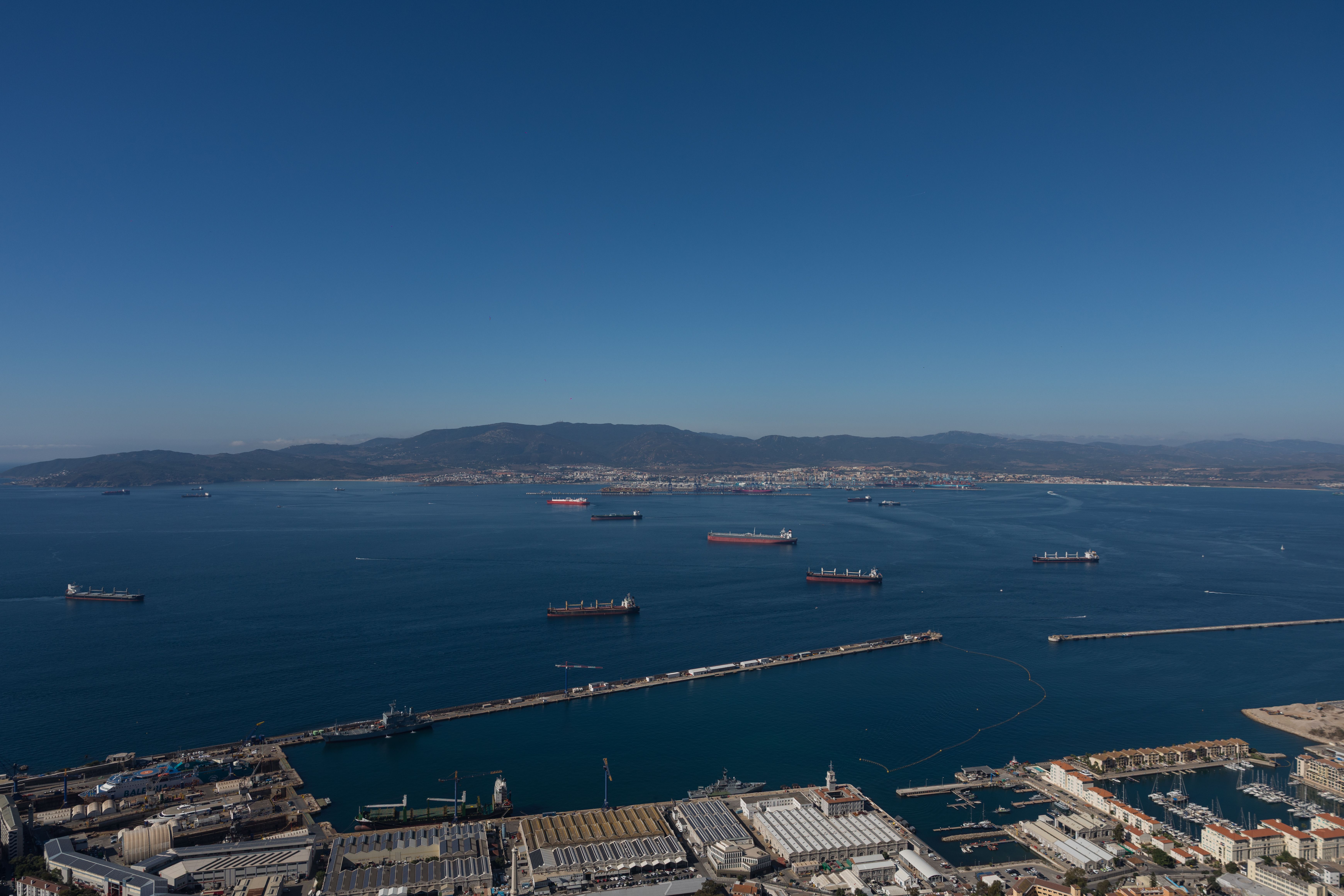 Fotomural Impresionante fotografía aérea del puerto de Gibraltar con barcos y montañas a lo lejos bajo un cielo azul despejado.