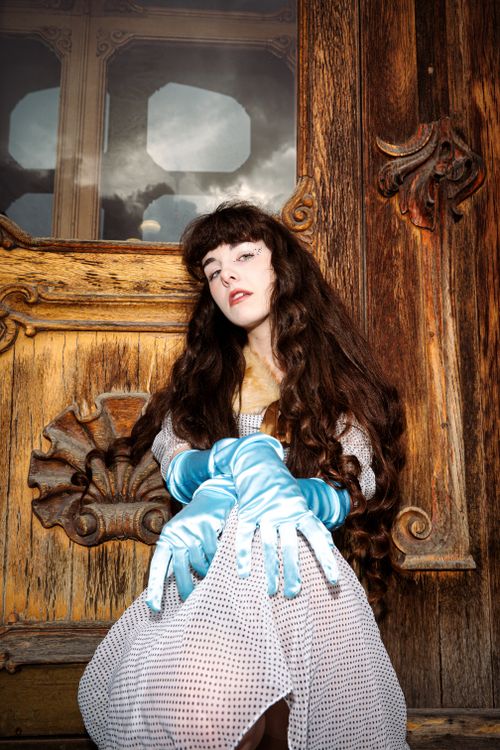 Free A woman with long hair and gloves poses elegantly by an ornate wooden door. Stock Photo
