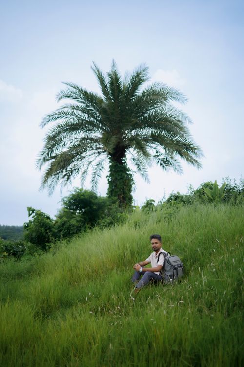 Free Man sits with backpack under a palm tree in a lush green landscape, enjoying nature. Stock Photo