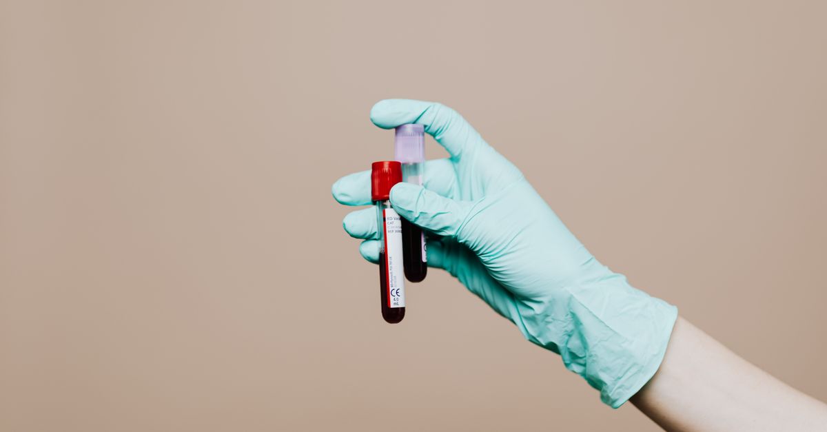 Close-up of a gloved hand holding blood test tubes against neutral background.
