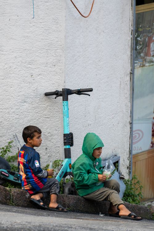 Free Two children seated next to a scooter on an urban street, wearing casual clothing. Stock Photo