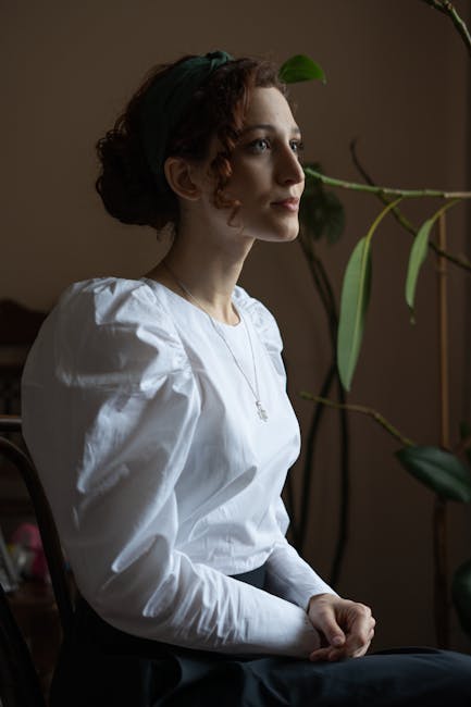 A woman with curly hair sits indoors, deep in thought, wearing a white blouse.