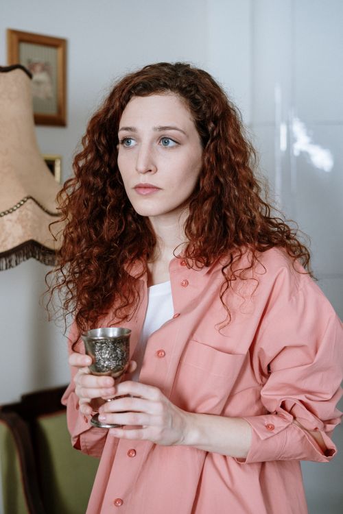 Free Young woman with red curly hair holding a goblet, standing indoors, wearing a pink shirt. Stock Photo
