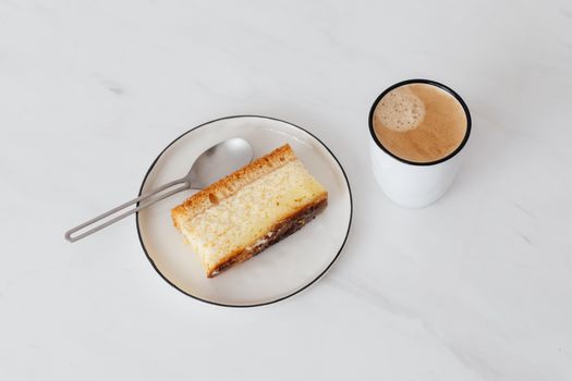 From above of cup of hot coffee near white plate with delicious piece of cake placed on white marble tabletop