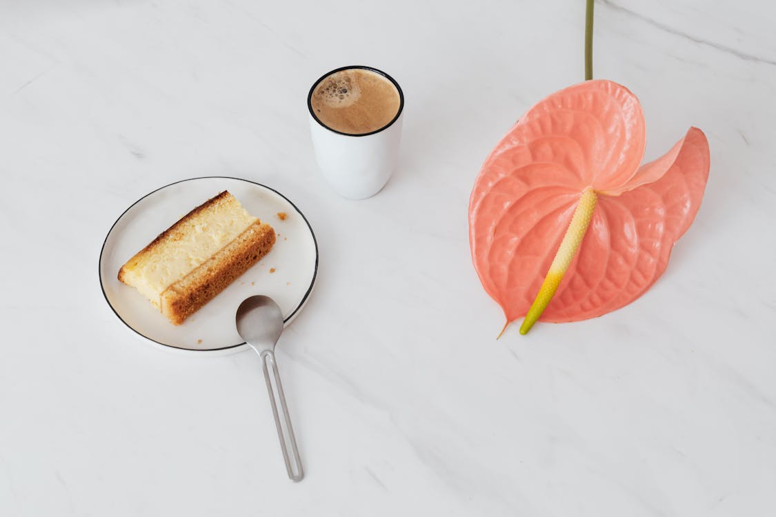 Free From above of pink Anthurium placed near ceramic cup of coffee and  fresh sweet dessert on ceramic plate with spoon on marble surface Stock Photo