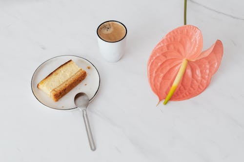 Free From above of pink Anthurium placed near ceramic cup of coffee and  fresh sweet dessert on ceramic plate with spoon on marble surface Stock Photo