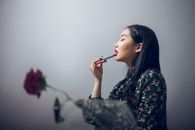 一位穿着时髦的亚洲女子站在花瓶旁，侧身对着玫瑰涂口红