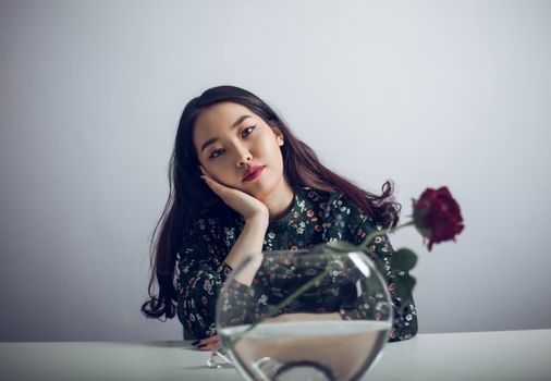 一位身穿印花连衣裙的年轻亚洲女子坐在桌旁，手捧一个插着芳香玫瑰的玻璃花瓶