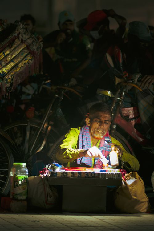 Free Elderly street vendor sells goods at night, illuminated by a small lamp. Stock Photo