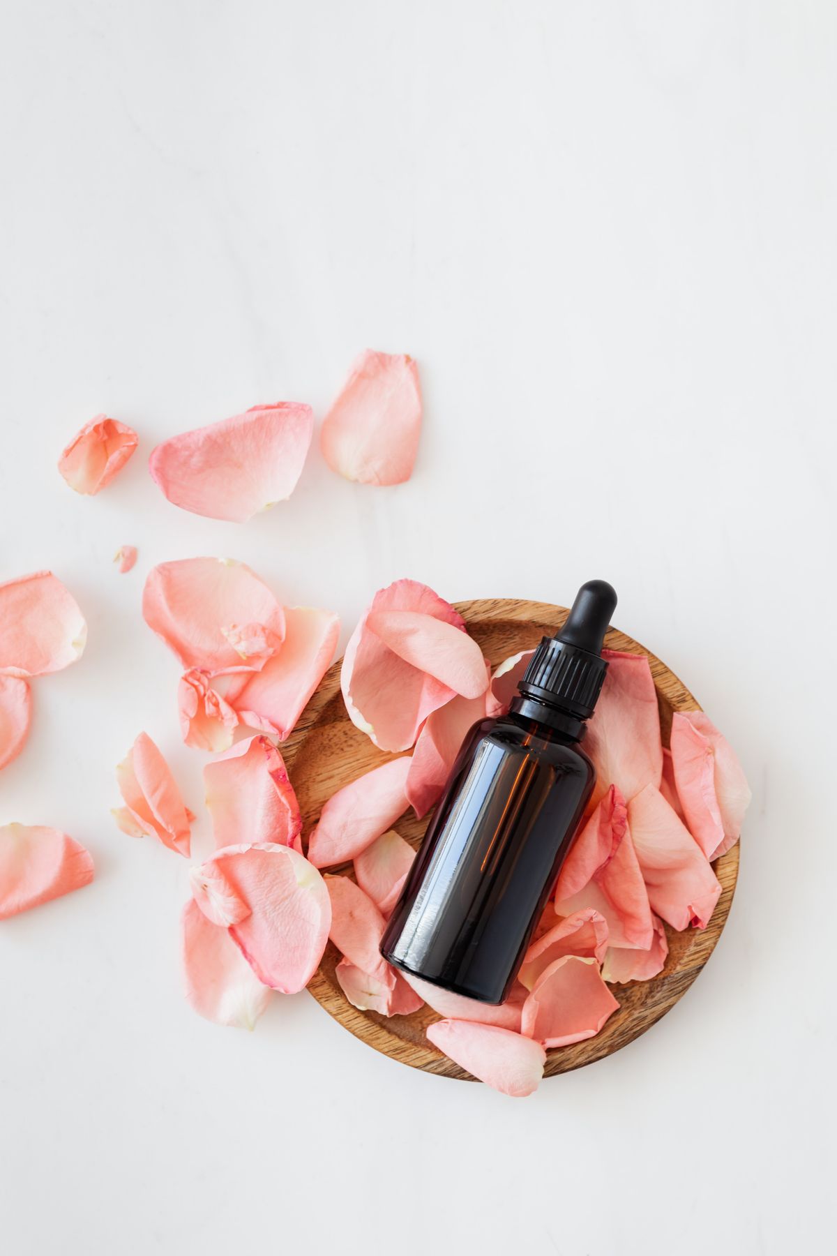 Brown glass dropper bottle with pale pink rose petals arranged on a round wooden plate against a white background