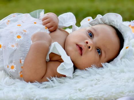 A cute baby lying outdoors on a fluffy blanket, dressed in a daisy-patterned summer outfit.