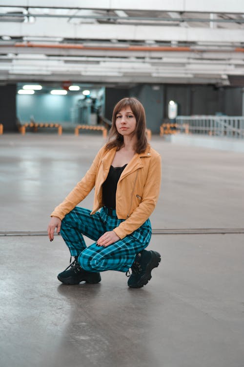 Full body young female in trendy jacket and trousers sitting on floor in industrial parking