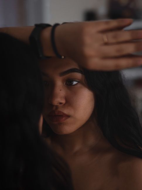 A contemplative woman gazes at her reflection in a dimly lit room, exploring self-doubt and identity.