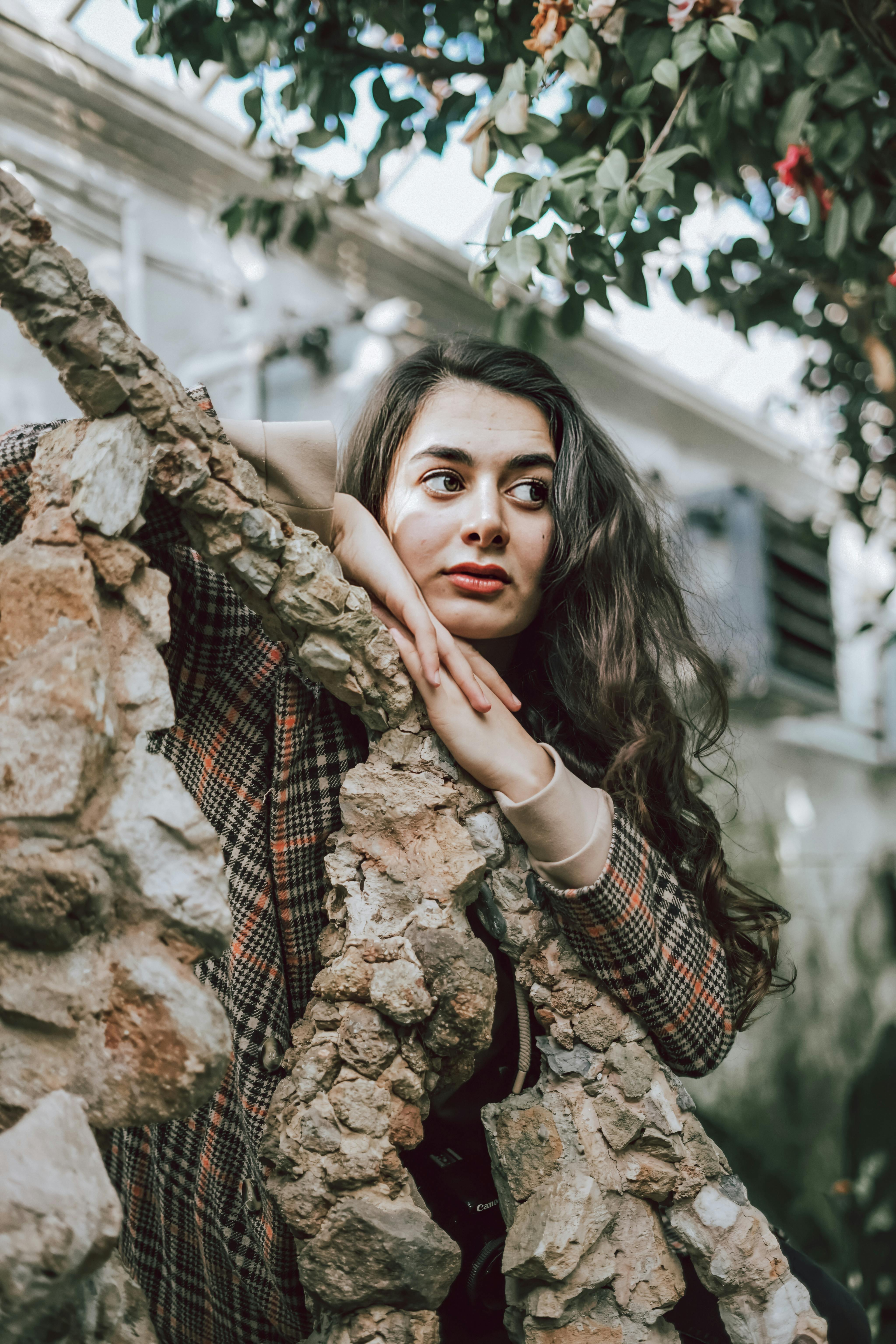 Dreaming brunette in checkered coat leaning on wooden trunk and looking away in hothouse