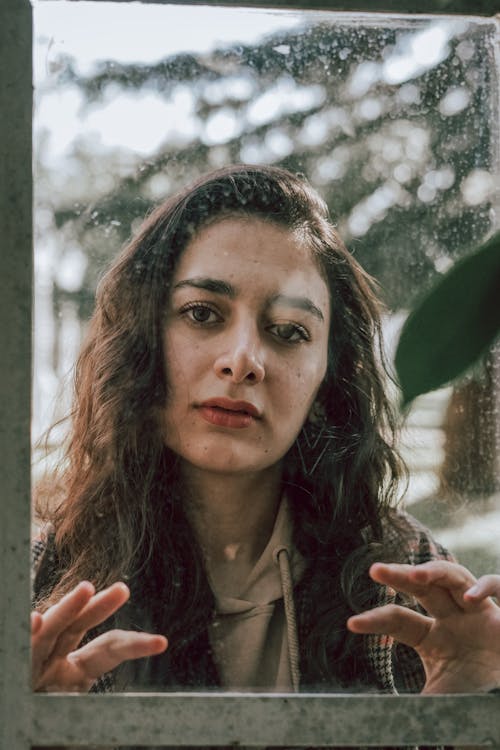 Melancholic brunette touching glass of window looking through it at camera against greenery