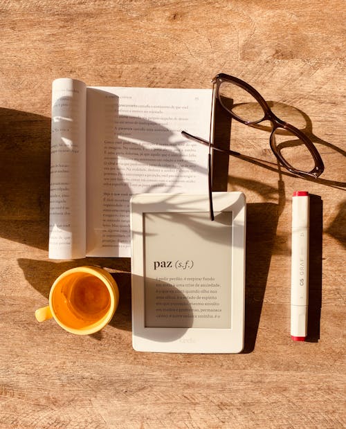 From above composition of e book and paper book on wooden table with cup and glasses in sunlight