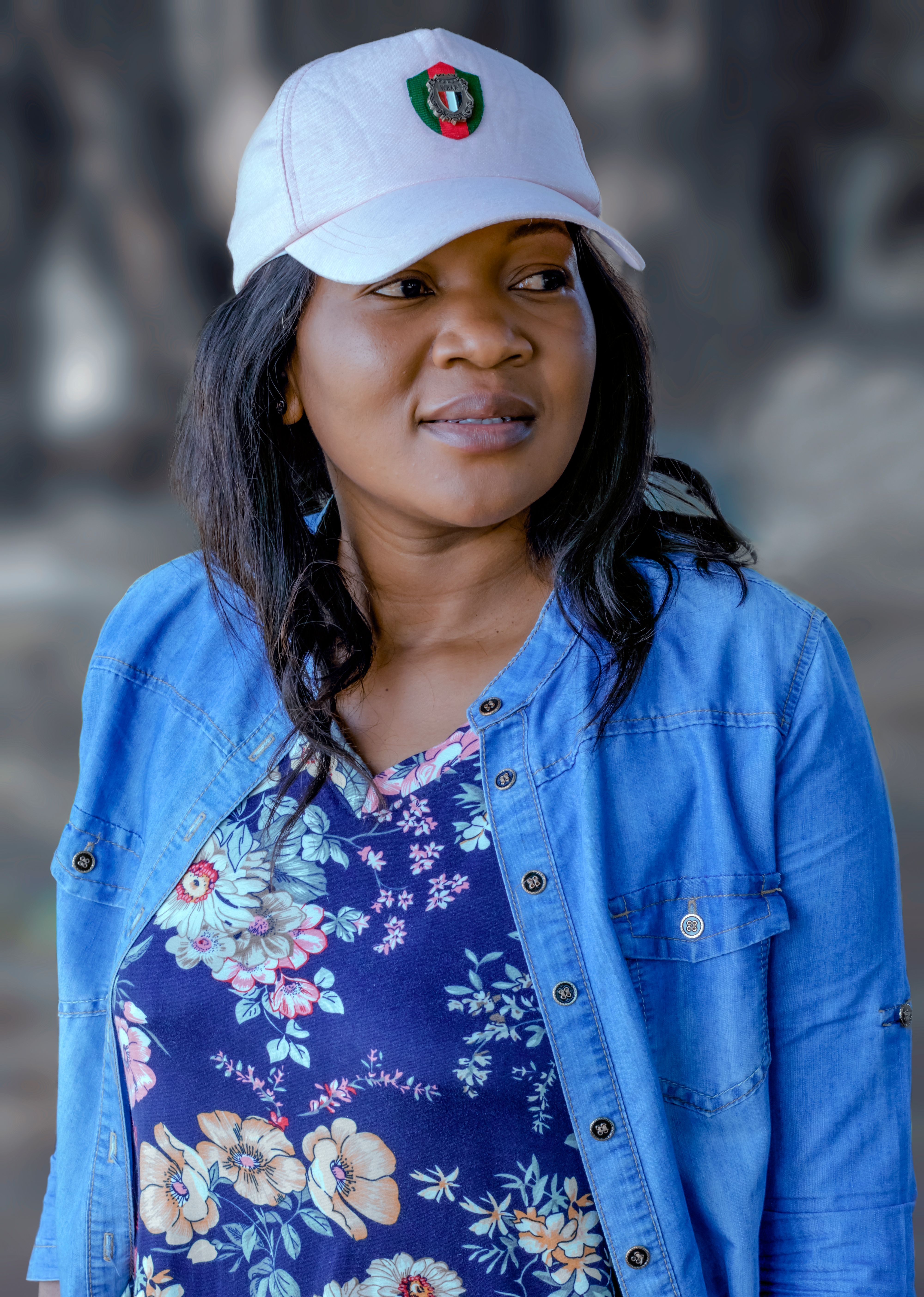 Portrait of a woman in a colorful floral top and denim jacket outdoors, looking away with a cap on.