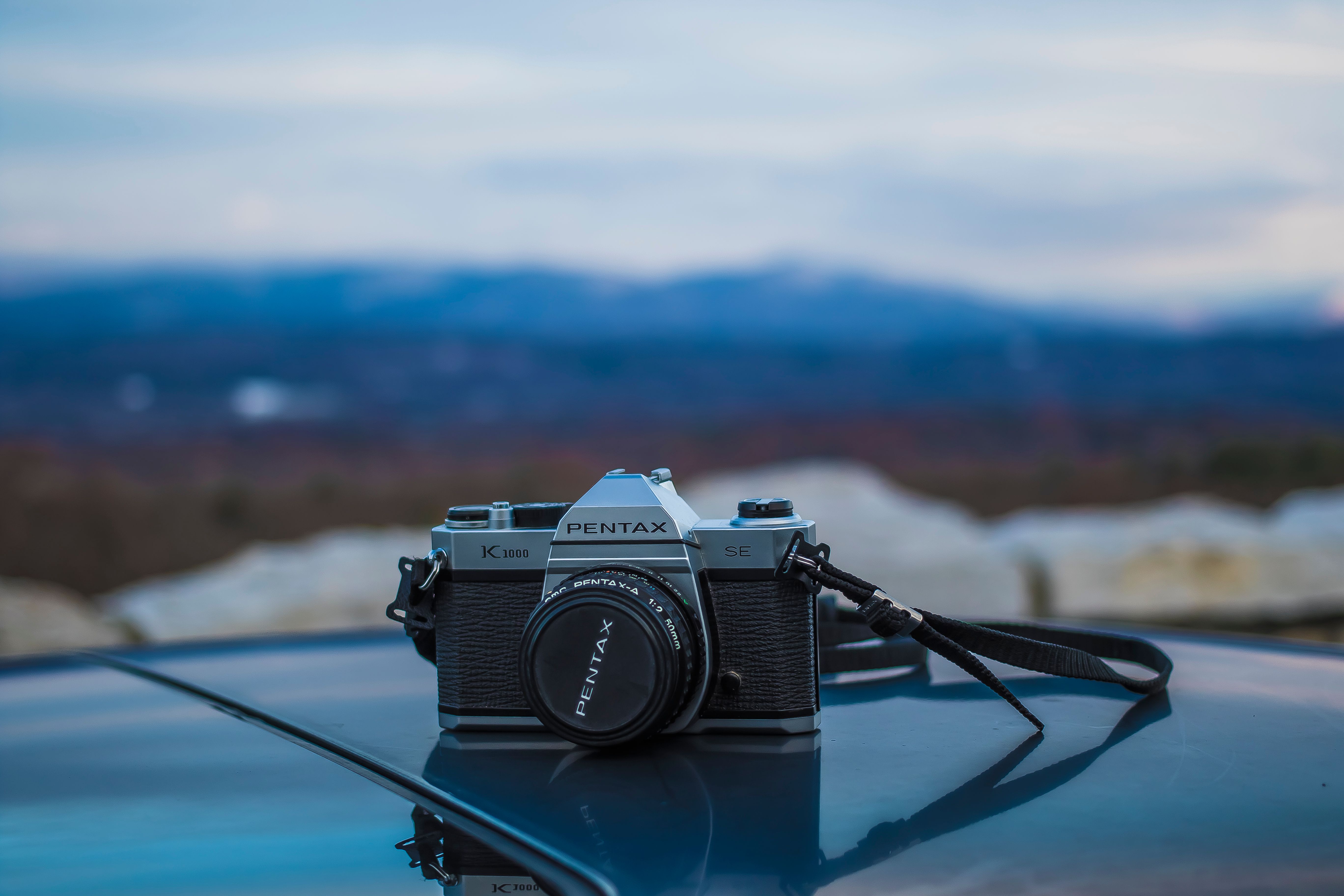 A classic Pentax camera rests on a car roof against a stunning mountain backdrop.