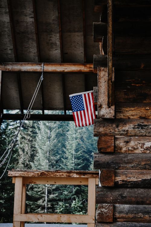 Free A rustic wooden cabin with an American flag in a forest setting, showcasing vintage architecture and natural surroundings. Stock Photo