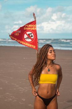 Cool slender woman with long braided hair standing on ocean beach with flag waving on background