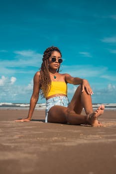 Full body young ethnic woman in shorts and top sitting confidently on sandy coast looking away