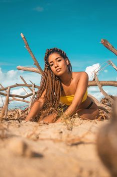 Slim young provocative ethnic female in swimsuit with dreadlocks sitting on sandy seashore with pointed tree twigs and looking at camera under blue sky in summer during vacation