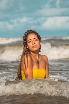 Young tattooed sun tanned ethnic female tourist with pendant and dreadlocks sitting in wavy sea with foamy water while looking at camera under cloudy sky