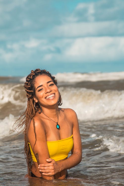 From above of positive slim ethnic female traveler in bright swimsuit with pendant looking at camera while enjoying foamy sea under cloudy sky during vacation