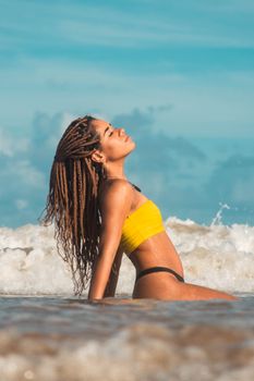 Side view of fit young provocative ethnic female with dreadlocks in swimwear sitting leaned on hands in wavy sea with closed eyes while sunbathing under sky in summer