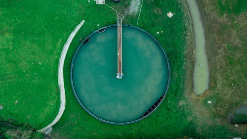 Aerial view of round shaped water treatment facility with rectangular metal construction surrounded by colorful green grass and narrow water stream in countryside