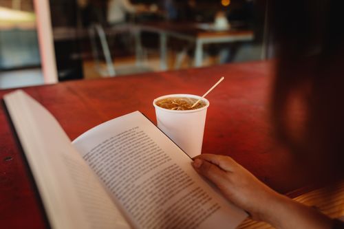Free A person enjoys a leisurely reading session with an iced drink on a warm tabletop. Stock Photo