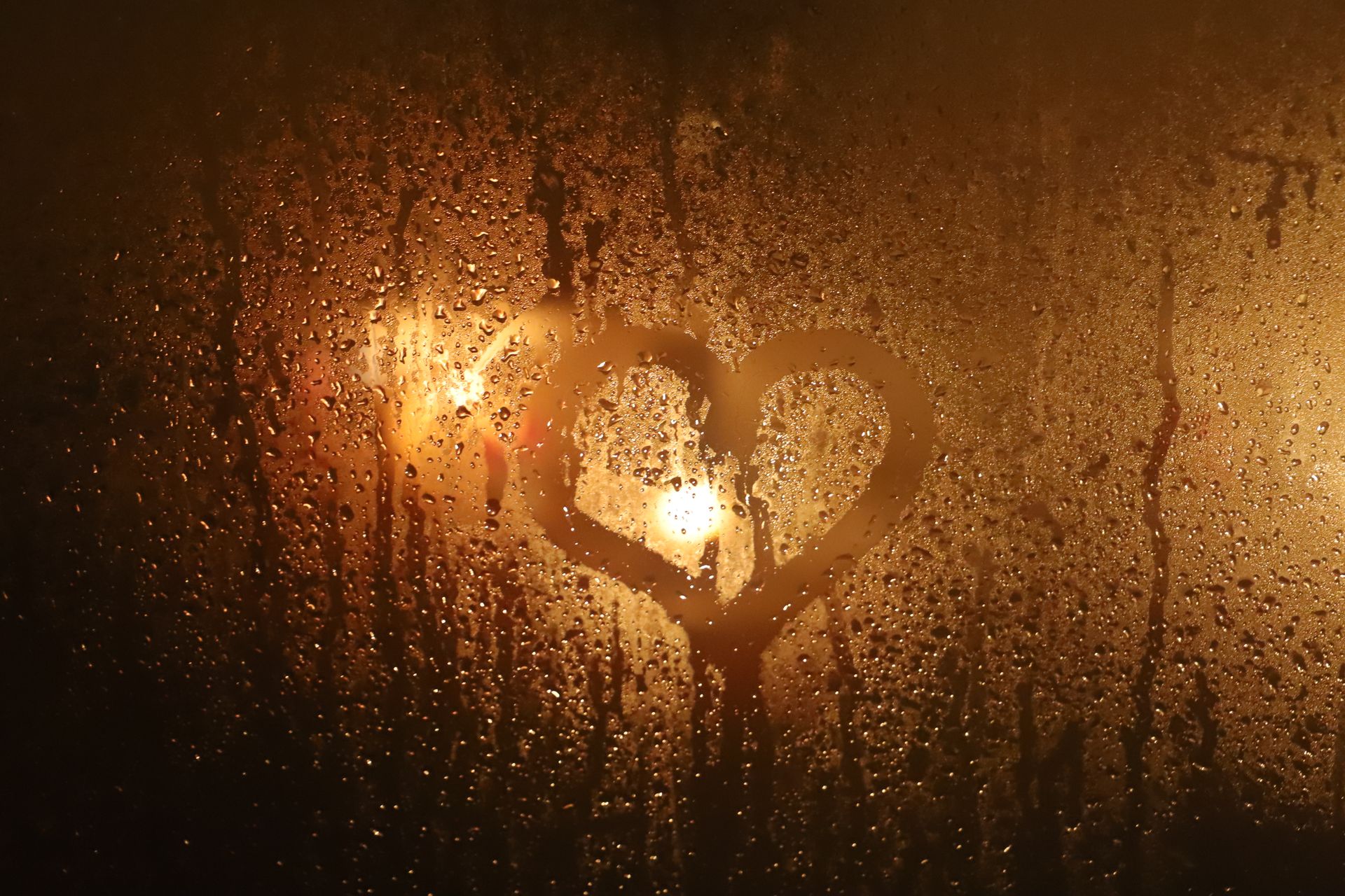 Symbol of heart on wet glass window with small drips and water fluids behind glowing lamps illuminating surface at night in rainy weather
