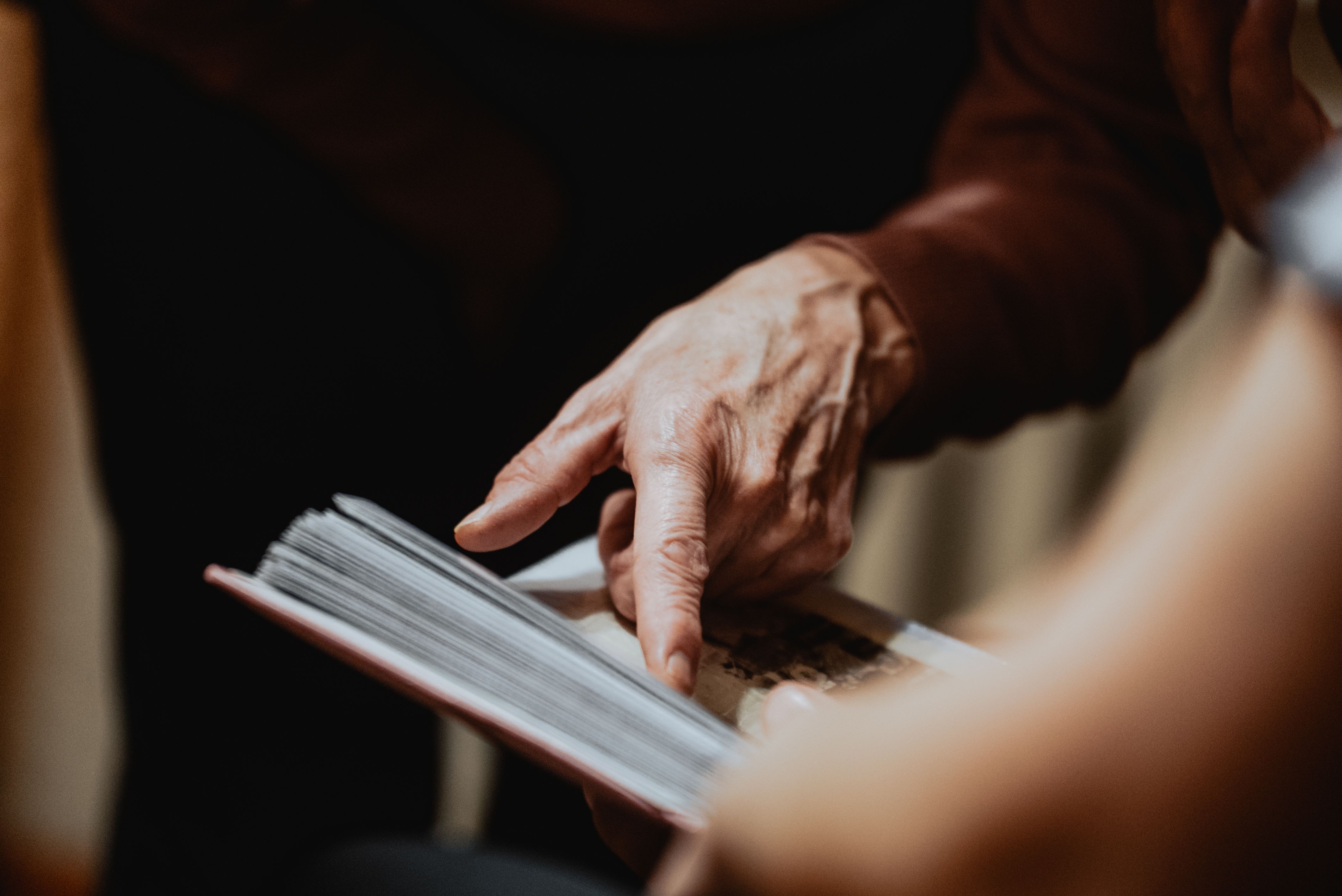 Elderly hand pointing at a page in an open book, suggesting detail and interaction.