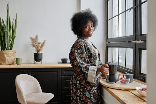 Smiling woman in a robe stands by the window making coffee, creating a cozy morning vibe.