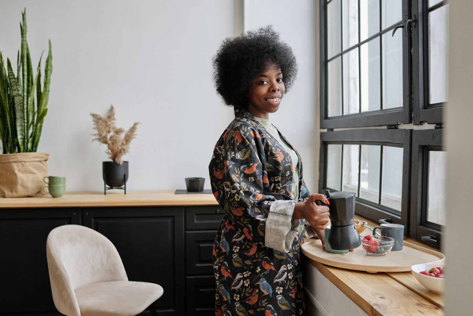 Smiling woman in a patterned robe making coffee by a large window in a bright kitchen