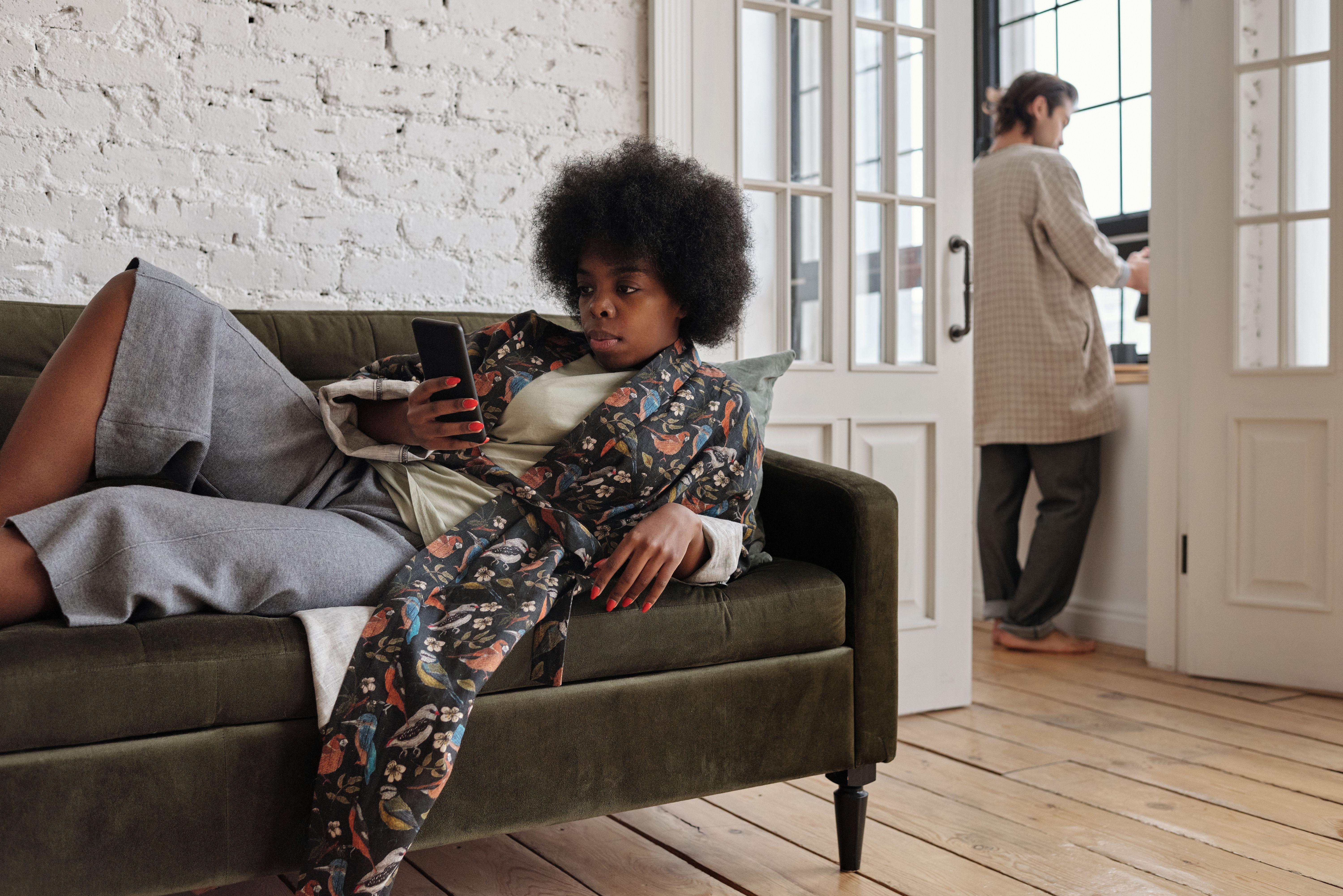 African American woman lounging on a sofa, texting, while man stands by window.